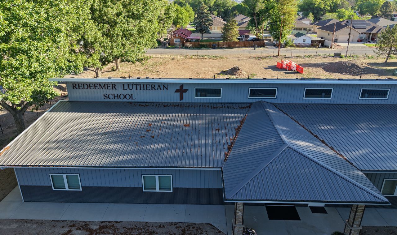 Aerial view of Redeemer Lutheran School building with a gray metal roof, large windows, and trees and houses visible in the background.